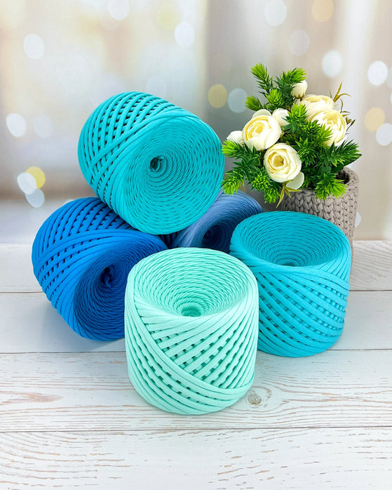 Three rolls of turquoise and blue yarn on a light wooden surface with a blurred floral background.