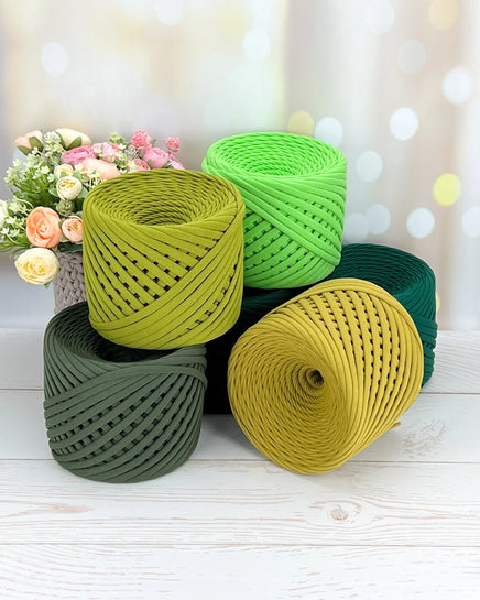 Five rolls of green and yellow yarn on a wooden surface with a blurred floral background.